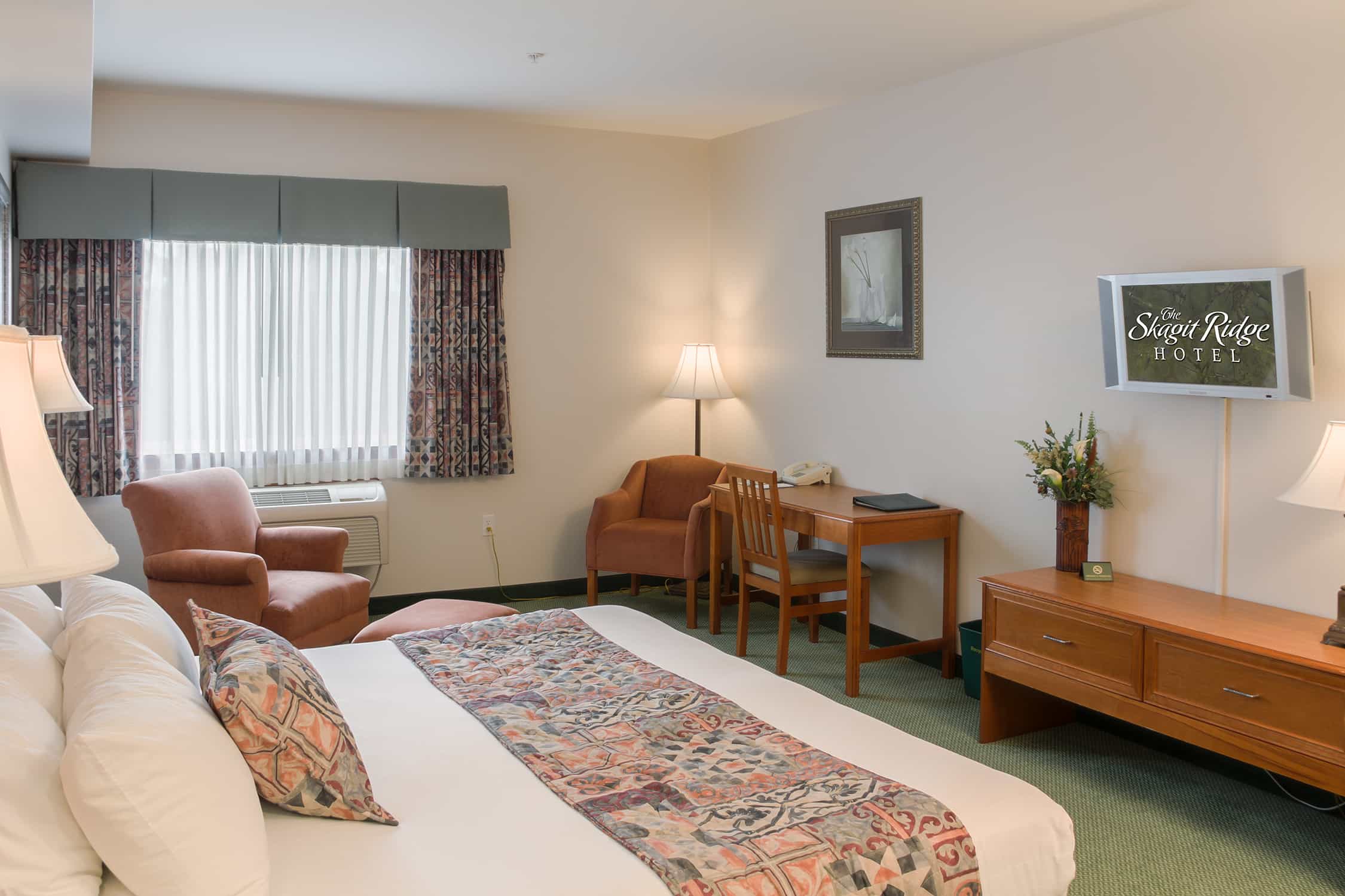 A Ridge Single King guest room, with one king bed dressed in white linen and a patterned runner, an armchair by the window, a writing desk and chair, a chest of drawers and a wall-mounted television