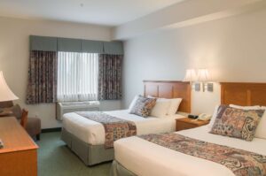 A Ridge Double Queen guest room, with two queen beds dressed in white linen and patterned runners, twin reading lamps above the headboards, a bedside telephone and a curtained window