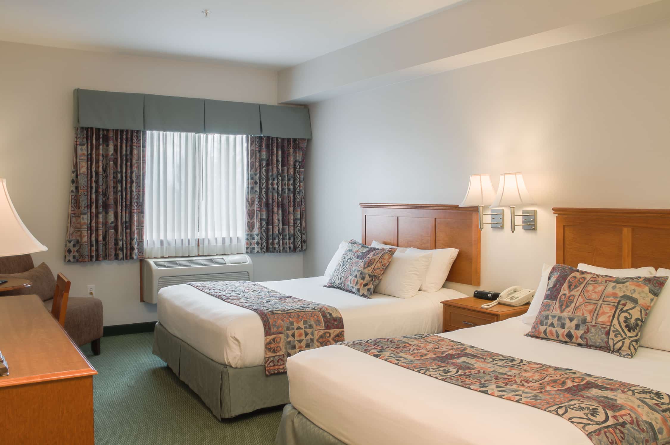 A Ridge Double Queen guest room, with two queen beds dressed in white linen and patterned runners, twin reading lamps above the headboards, a bedside telephone and a curtained window