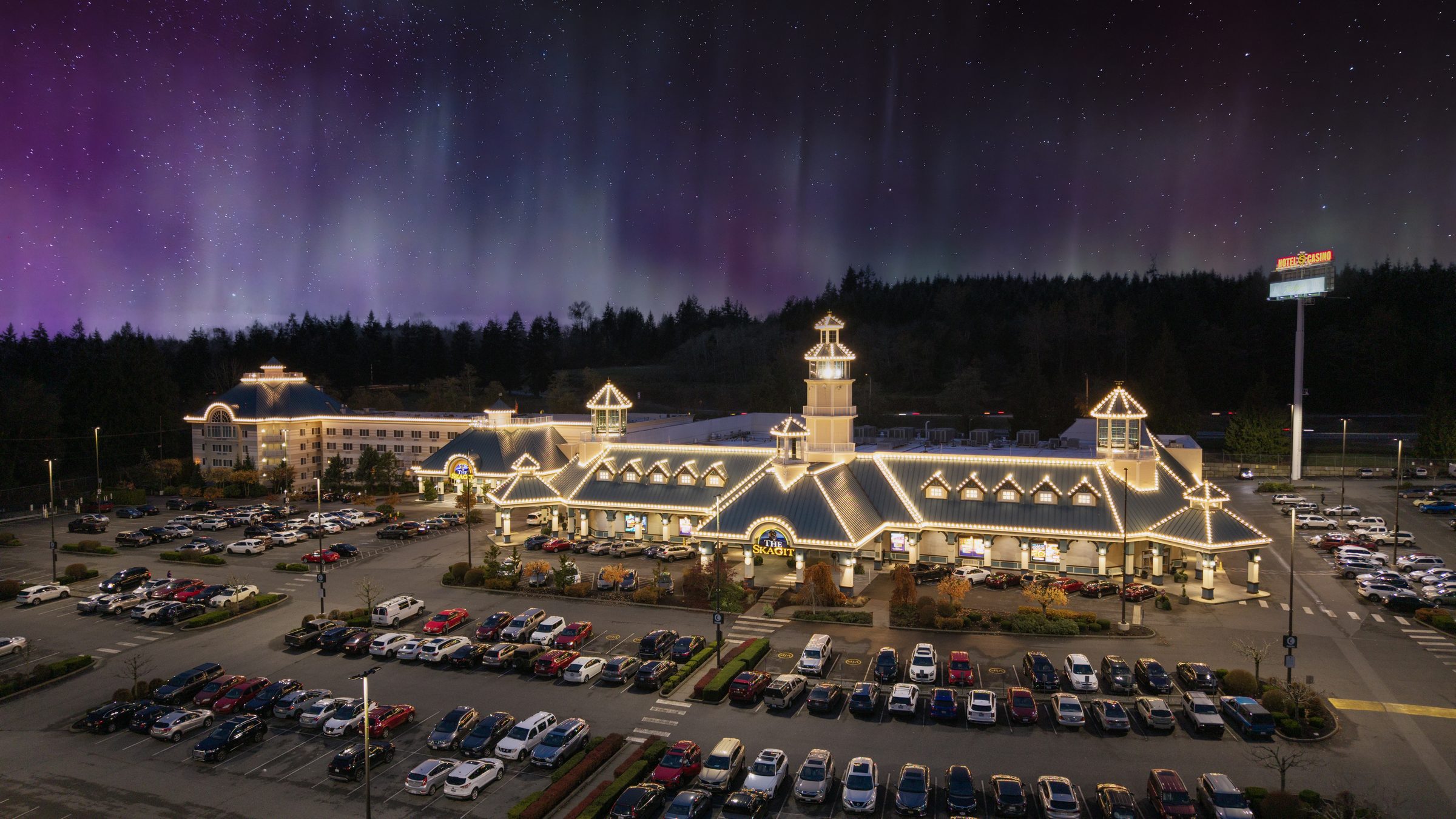 The northern lights in purple and green above The Skagit Casino Resort at night, the building glowing along its rooflines